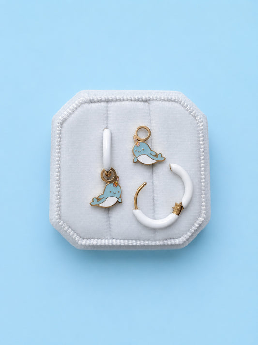 Pair of huggie hoops with narwhal charms on a white cushion against a light blue background