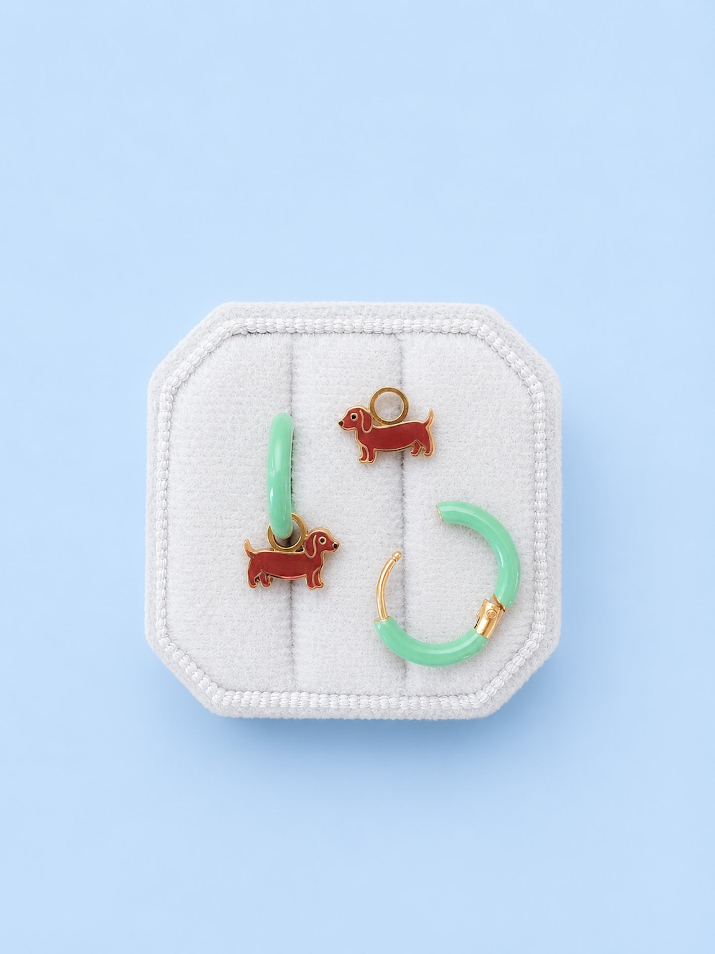 A set of earrings displayed on a white fabric jewelry stand against a light blue background. The set includes two small brown dachshund dog charms, a mint green bar, and a matching mint green hoop with gold ear wires.