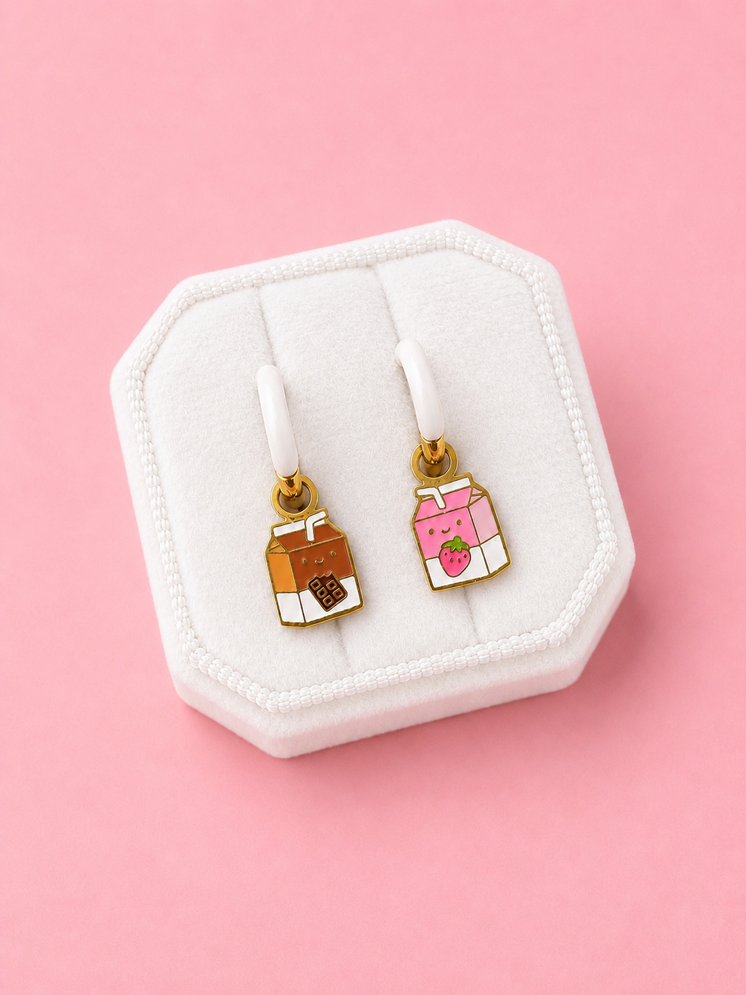 A pair of small enamel drop earrings displayed on a white jewelry stand against a pink background. One earring features a brown milk carton with a chocolate heart, and the other features a pink milk carton with a strawberry.