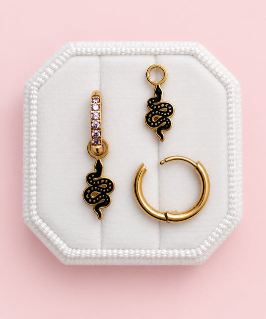 Three gold-toned earrings displayed on a white octagonal jewelry box against a soft pink background. The set includes two snake-shaped drop earrings with black enamel detailing, one with small purple stones, and one large gold hoop earring.