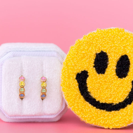 Colorful earrings in a white case next to a smiley face sponge on a pink background