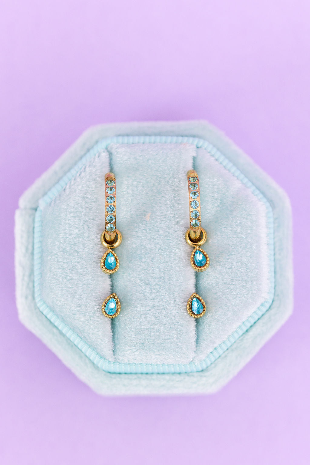 Pair of gold and turquoise earrings on a light blue hexagonal stand against a purple background