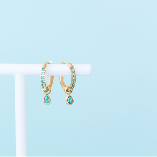 Gold hoop earrings with green gemstones on a white stand against a light blue background