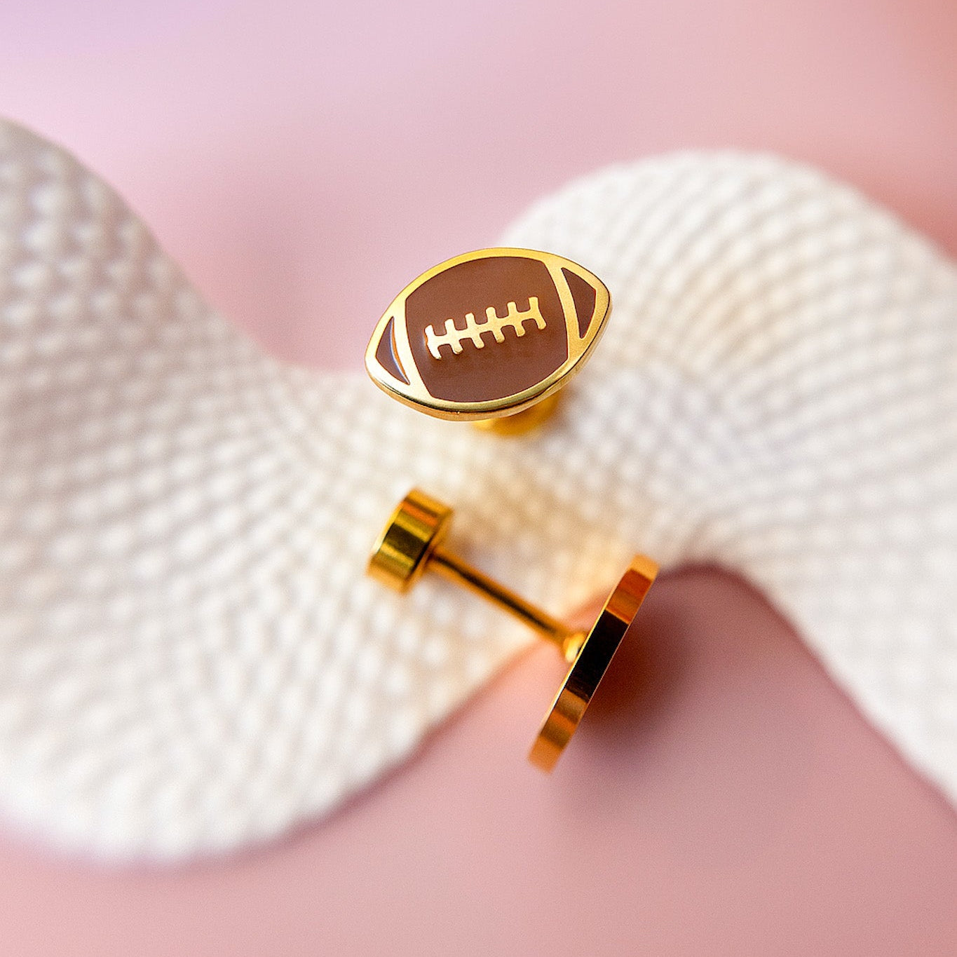 Gold football-shaped earrings on a textured white surface with a pink background