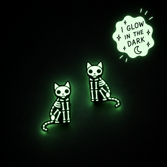 Two skeleton cat glow-in-the-dark earrings on a black background, with one pin featuring a speech bubble that reads 'I GLOW IN THE DARK' with a crescent moon.