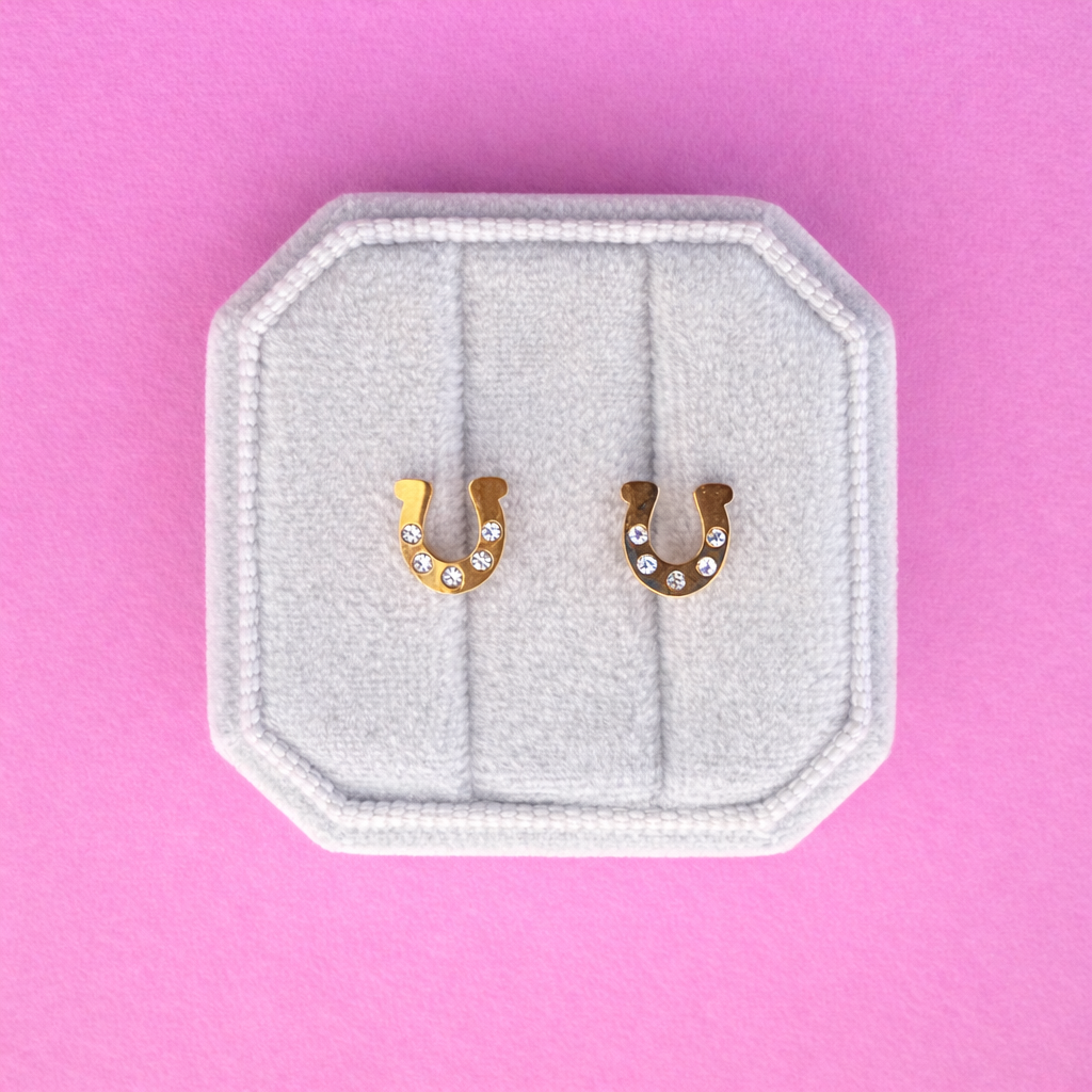 Horseshoe-shaped earrings with rhinestones on a white cushion against a pink background