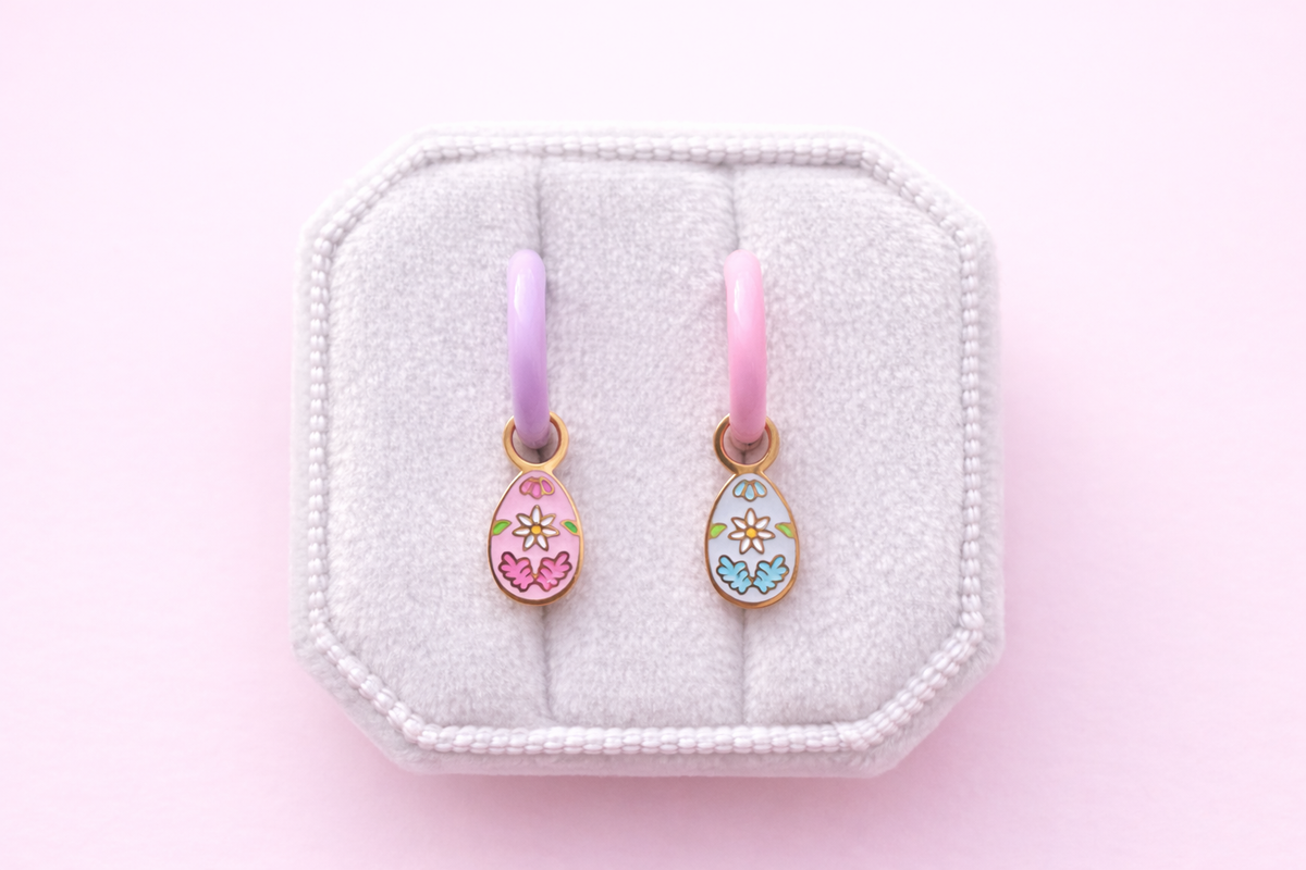 Floral egg charms 1 pink and 1 light blue. The pink one is on a lavender hoop and the light blue is on light pink hoop. Both are on a white jewelry cushion against a light pink background