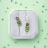 Pair of green milkshake-shaped charms on a white huggie hoop on a white cushion against a light green background with small green sprinkles