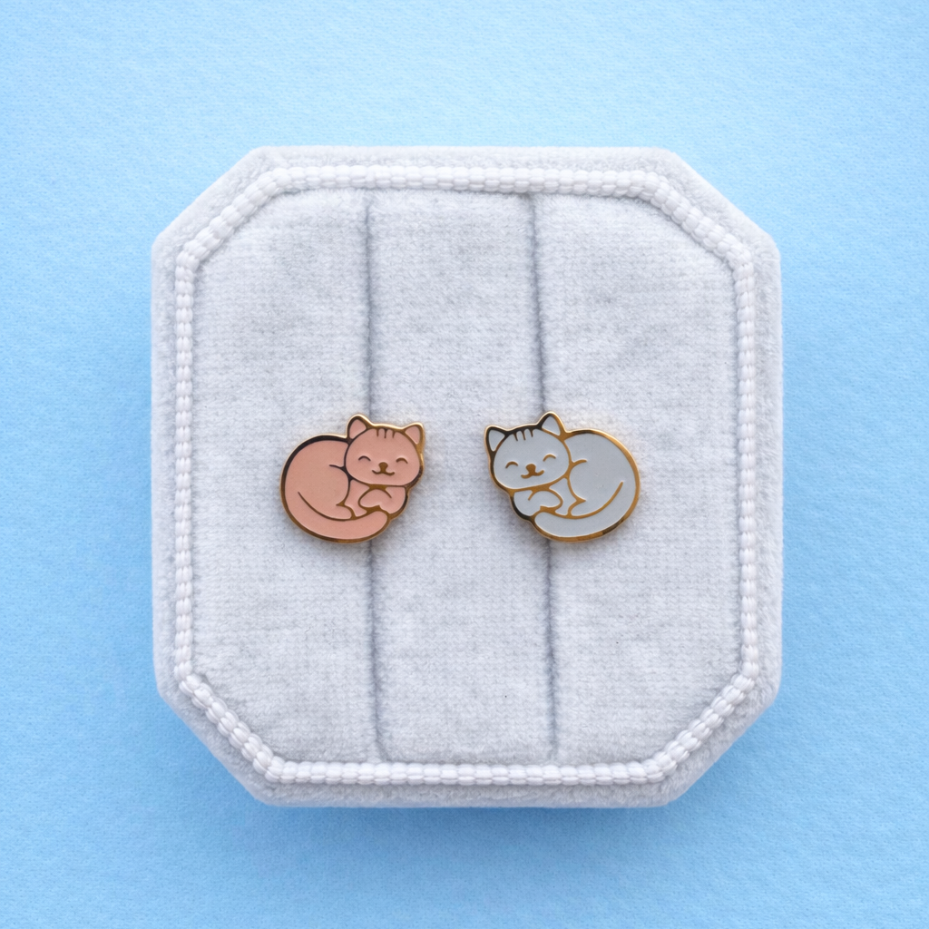 Tabby cat themed screw on flat back earrings with an orange cat and a light blue cat on a white jewelry box cushion against a light blue background