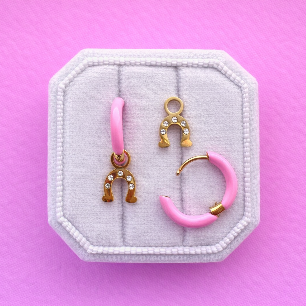 Two light pink huggie hoops with horseshoe charms with cz accents on a white jewerly box against a pink background