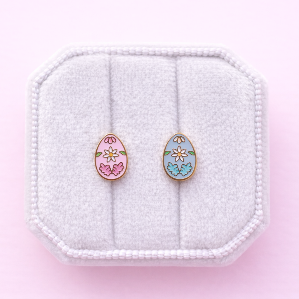 A set of floral egg stud earrings 1 pink and 1 light blue on a white jewelry box cushion against a light pink background