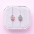 A set of floral egg stud earrings 1 pink and 1 light blue on a white jewelry box cushion against a light pink background