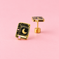 A pair of gold and black enamel earrings shaped like a book of spells, featuring a crescent moon and the word 'SPELLS', displayed on a solid pink background.