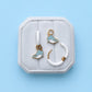 Pair of huggie hoops with narwhal charms on a white cushion against a light blue background
