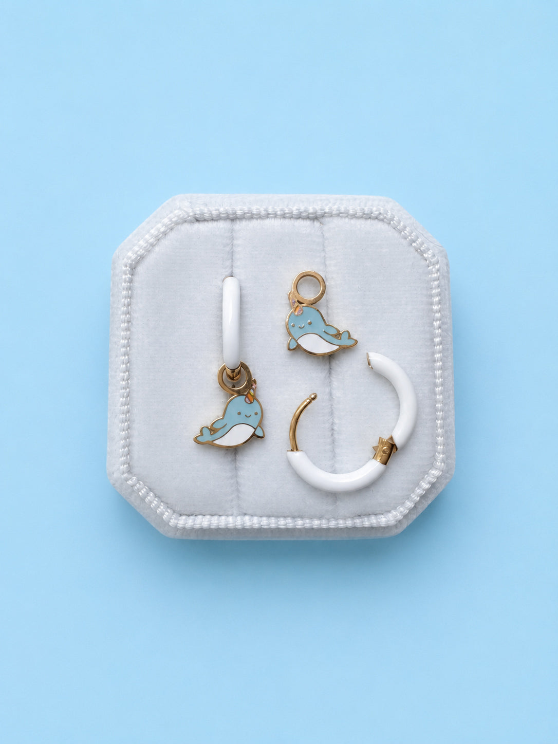 Pair of huggie hoops with narwhal charms on a white cushion against a light blue background