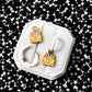 A pair of gold hoop earrings with colorful crayon box charms, displayed on a white octagonal jewelry stand against a black and white starry patterned background.