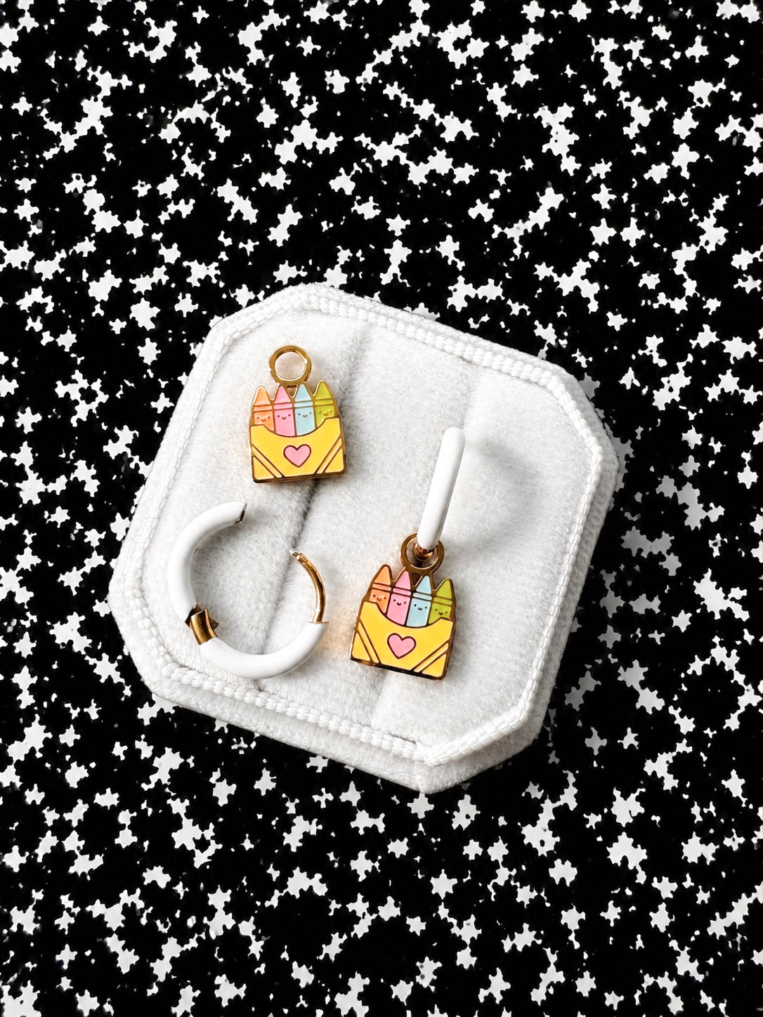 A pair of gold hoop earrings with colorful crayon box charms, displayed on a white octagonal jewelry stand against a black and white starry patterned background.