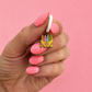 A hand with pink manicured nails holds a small enamel charm shaped like a yellow crayon box with four colorful crayons and a pink heart, against a solid pink background.