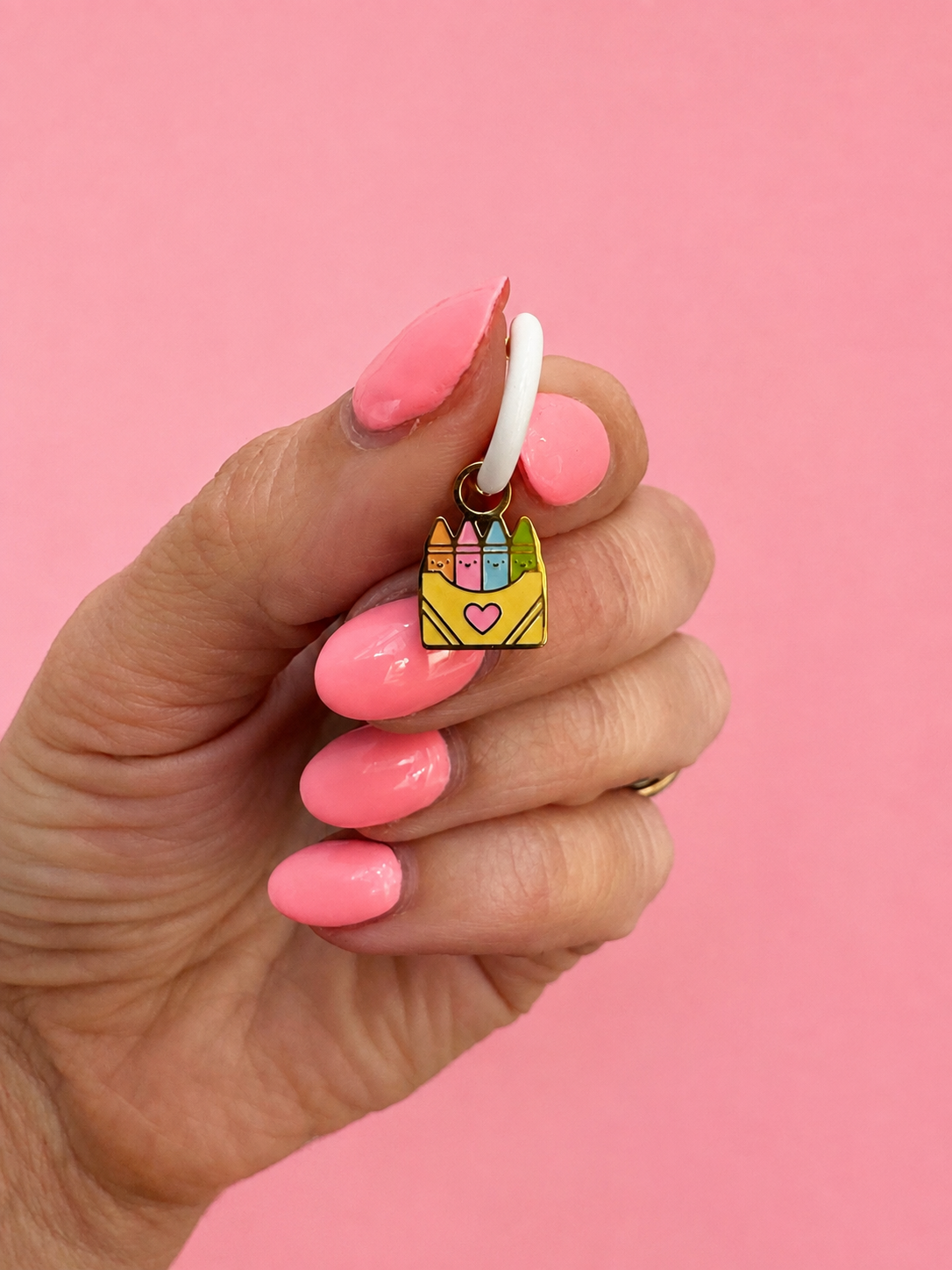 A hand with pink manicured nails holds a small enamel charm shaped like a yellow crayon box with four colorful crayons and a pink heart, against a solid pink background.