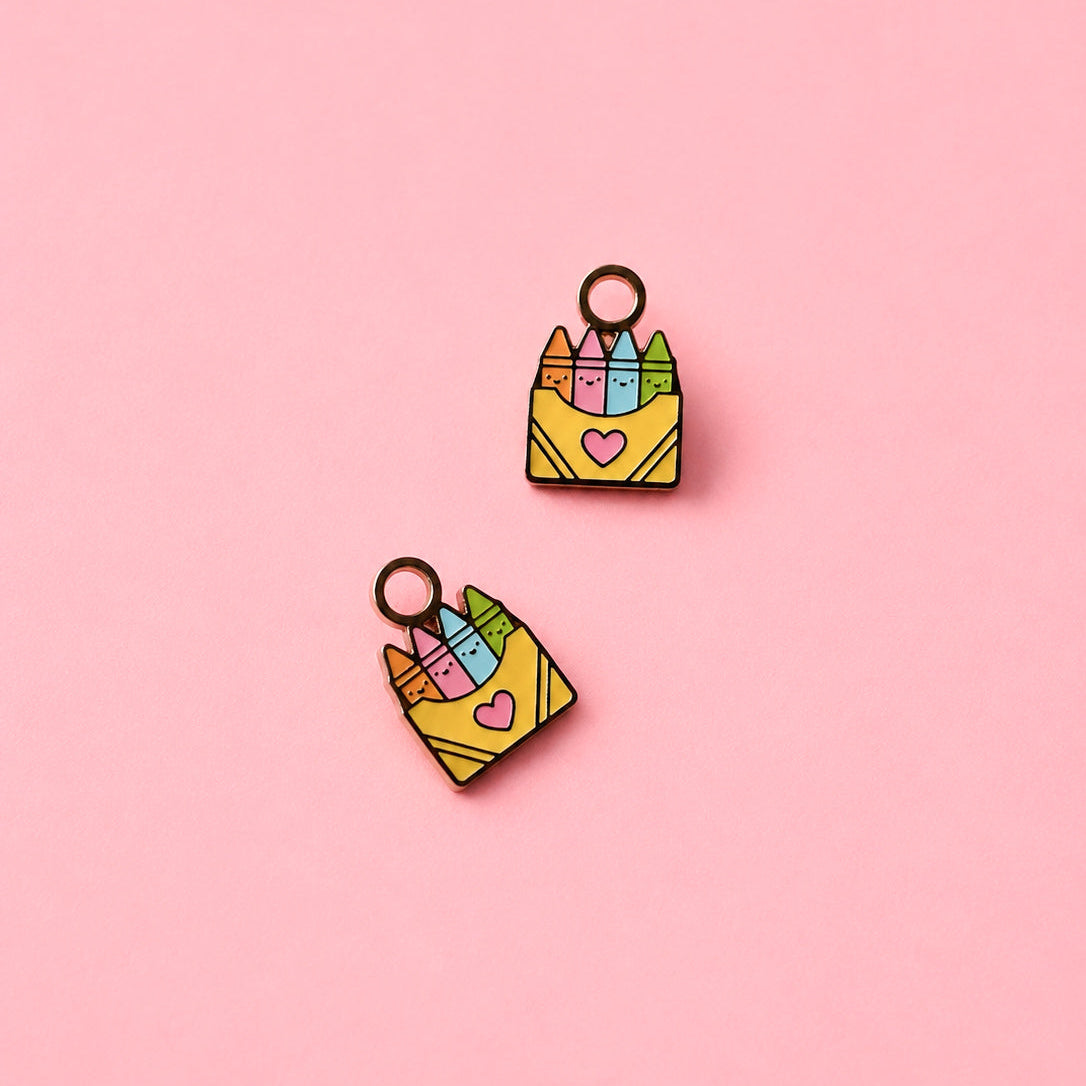 Two small enamel charms shaped like yellow crayon boxes filled with colorful crayons, shown on a solid pink background.
