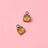 Two small enamel charms shaped like yellow crayon boxes filled with colorful crayons, shown on a solid pink background.