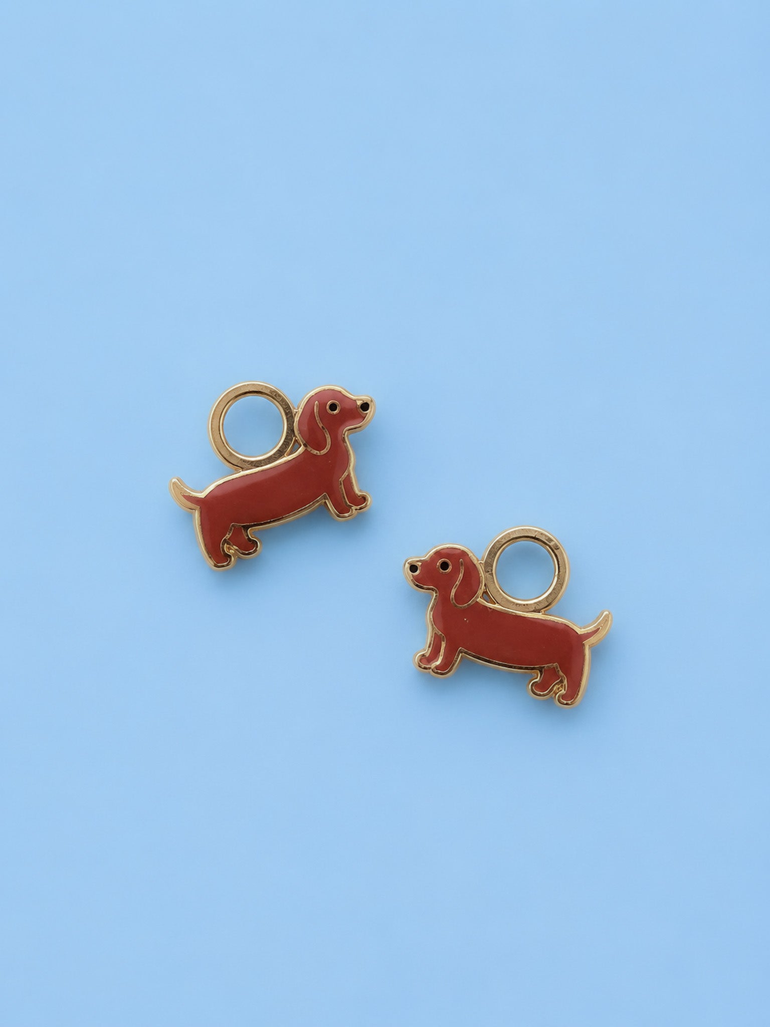 Two small dachshund-shaped jewelry charms with gold-tone metal outlines and loops, shown on a light blue background.