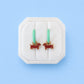 A pair of small hoop earrings with mint green hoops and red dachshund-shaped charms, displayed on a white octagonal jewelry stand against a light blue background.