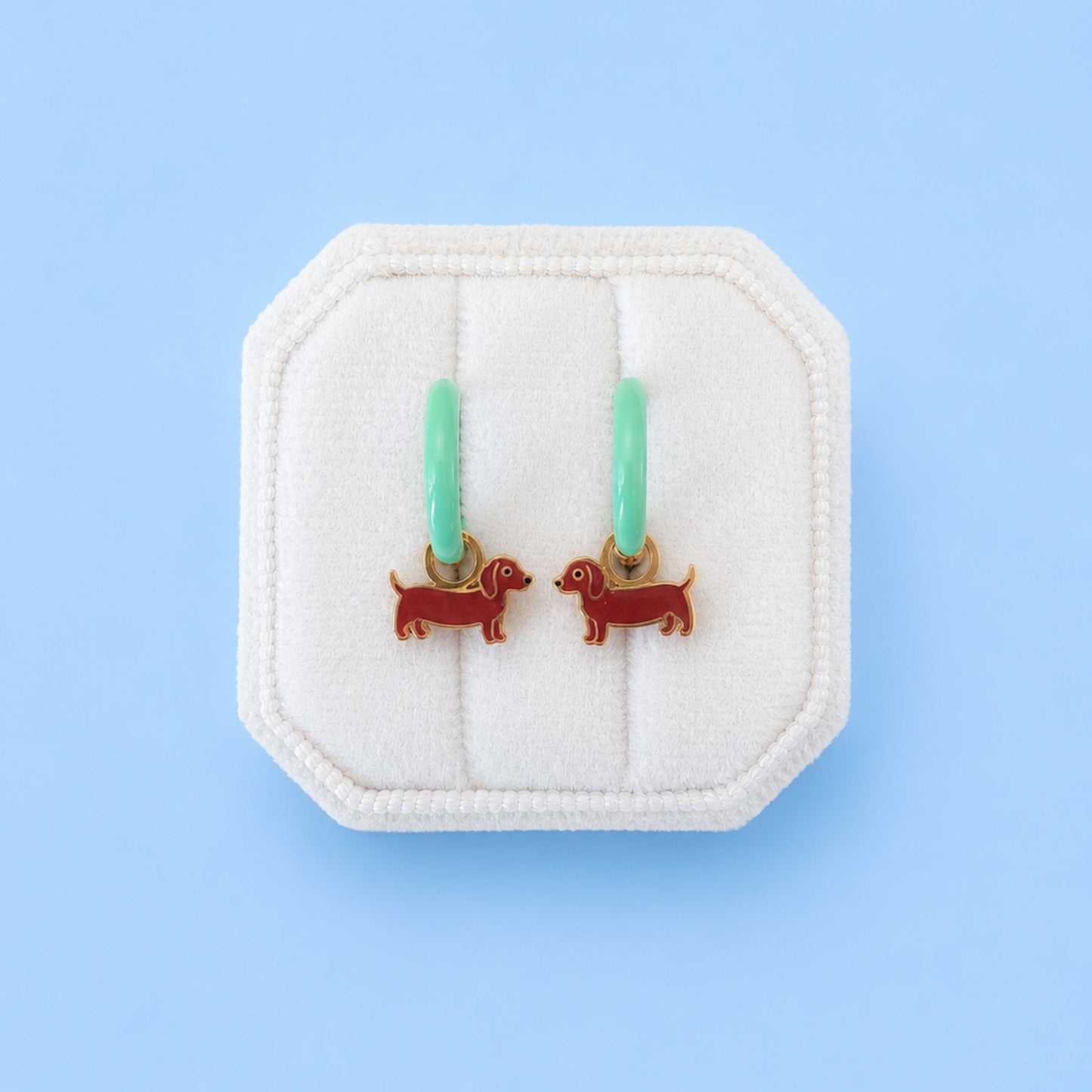 A pair of small hoop earrings with mint green hoops and red dachshund-shaped charms, displayed on a white octagonal jewelry stand against a light blue background.