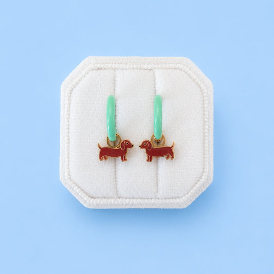 A pair of small hoop earrings with mint green hoops and red dachshund-shaped charms, displayed on a white octagonal jewelry stand against a light blue background.