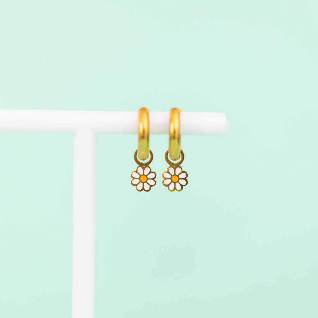 White daisy huggie charms on gold huggie hoops hanging on a white jewelry stand against a light green background