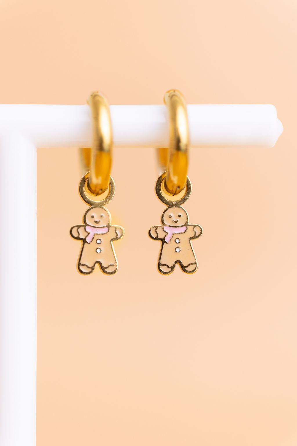 Gingerbread huggie charms on 8mm gold huggie hoops hanging on a white jewelry stand against a light peach background