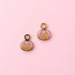 Two small pumpkin-shaped charms with gold-tone loops, pink glittery fill, and yellow jack-o'-lantern faces, arranged on a solid pink background.