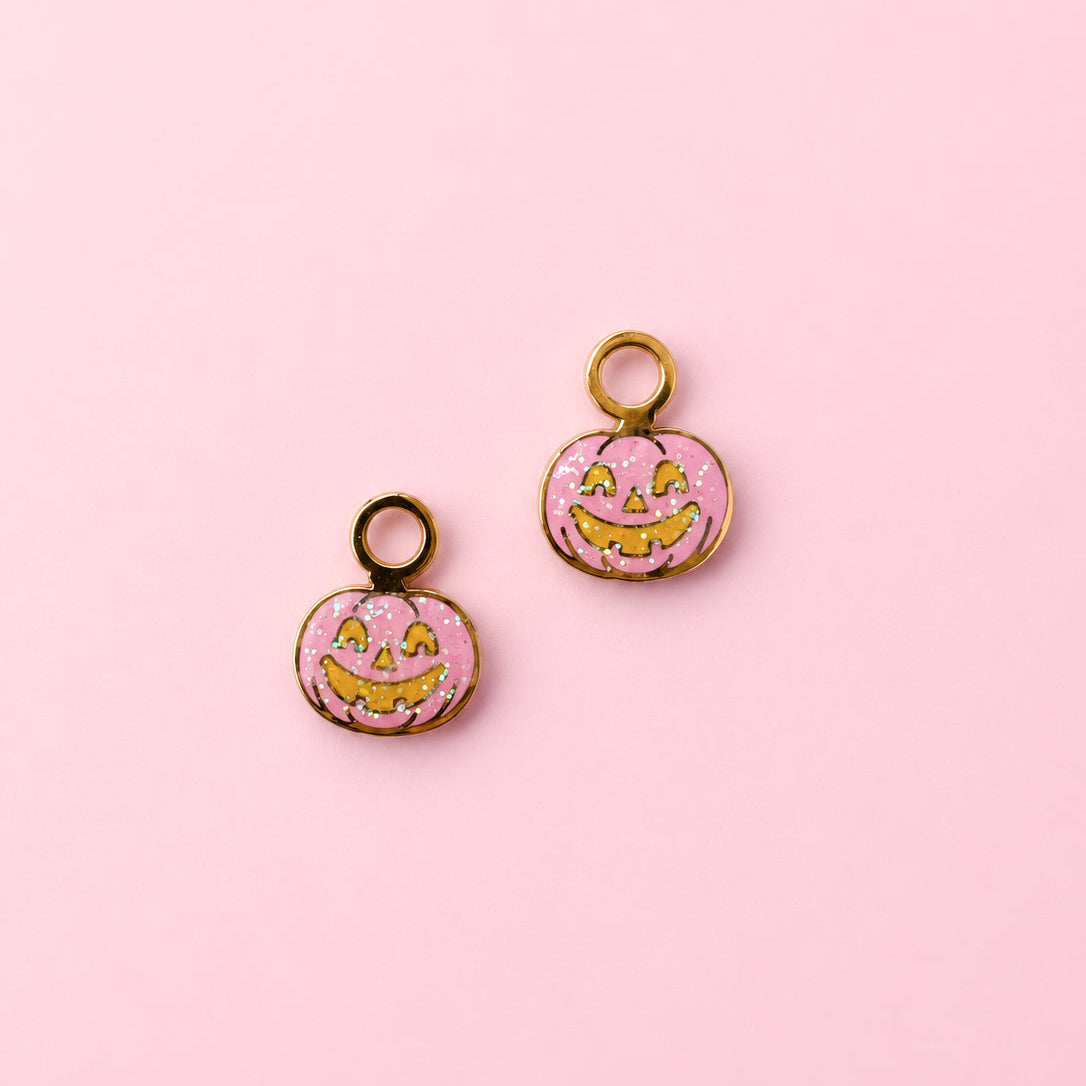 Two small pumpkin-shaped charms with gold-tone loops, pink glittery fill, and yellow jack-o'-lantern faces, arranged on a solid pink background.