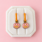 Two small pumpkin-shaped charms with oarnge-tone hoops, pink glittery fill, and yellow jack-o'-lantern faces, arranged on a solid pink background.