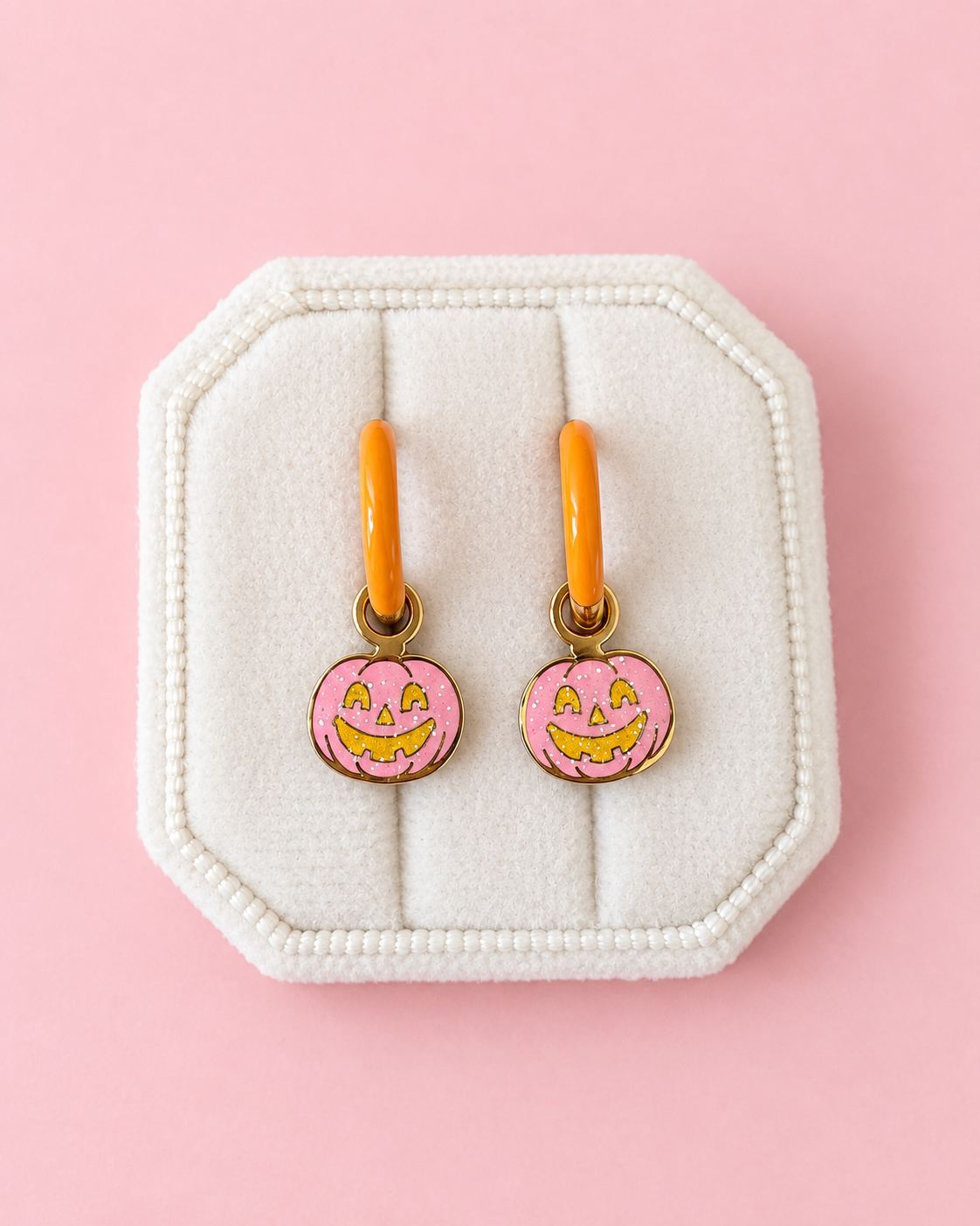 Two small pumpkin-shaped charms with oarnge-tone hoops, pink glittery fill, and yellow jack-o'-lantern faces, arranged on a solid pink background.