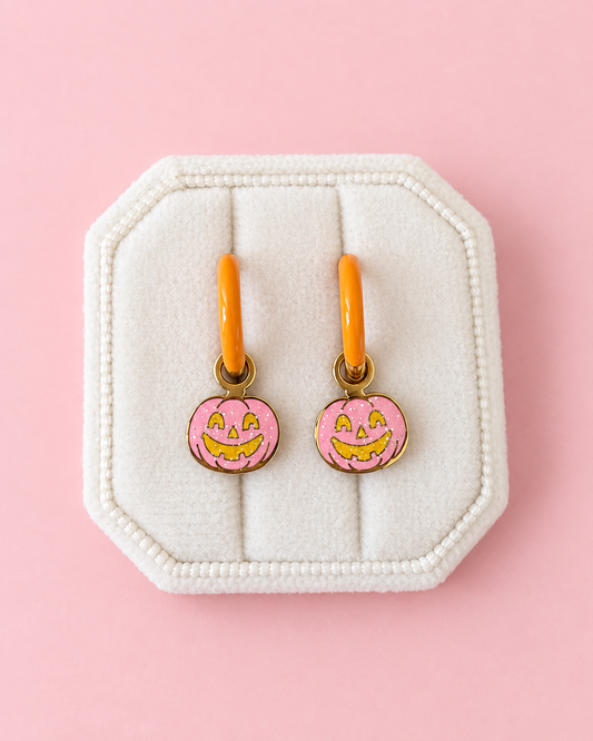 Two small pumpkin-shaped charms with oarnge-tone hoops, pink glittery fill, and yellow jack-o'-lantern faces, arranged on a solid pink background.
