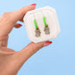Hand holding Koala huggie charms on green huggie hoops with white jewelry box with blue background