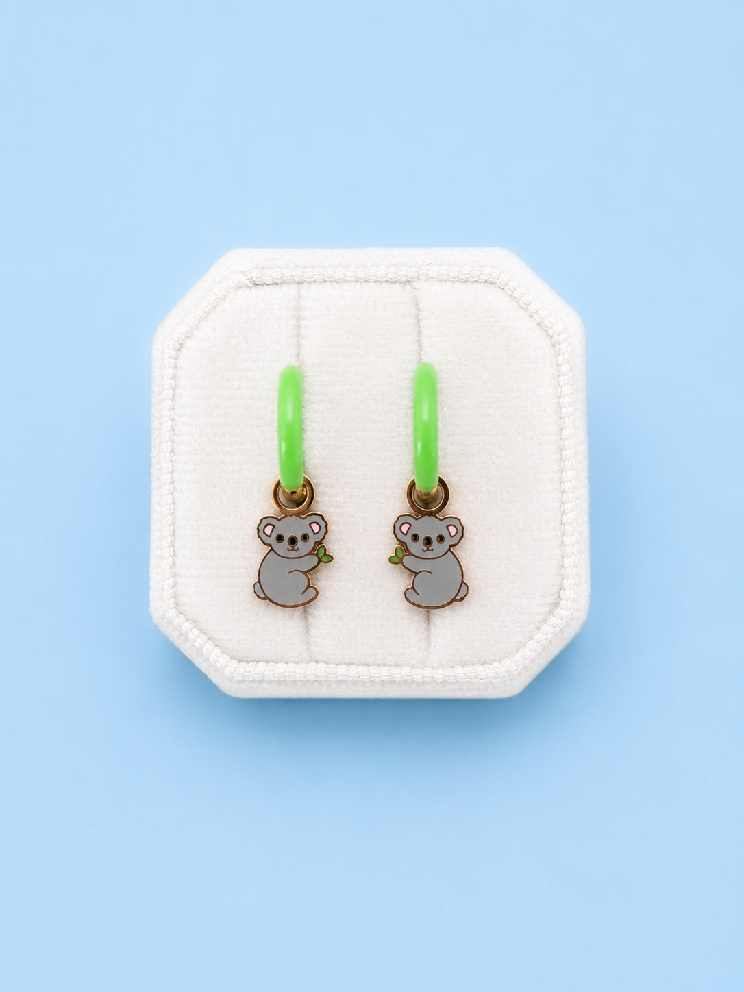 Koala huggie charms on green huggie hoops on white jewelry box with blue background