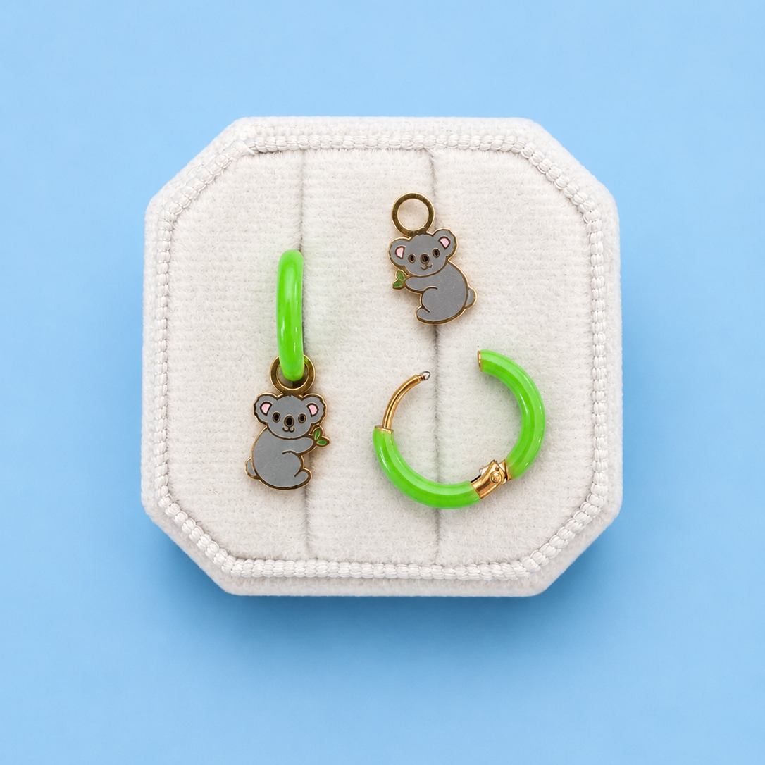 Koala huggie charms on green huggie hoops on white jewelry box with blue background