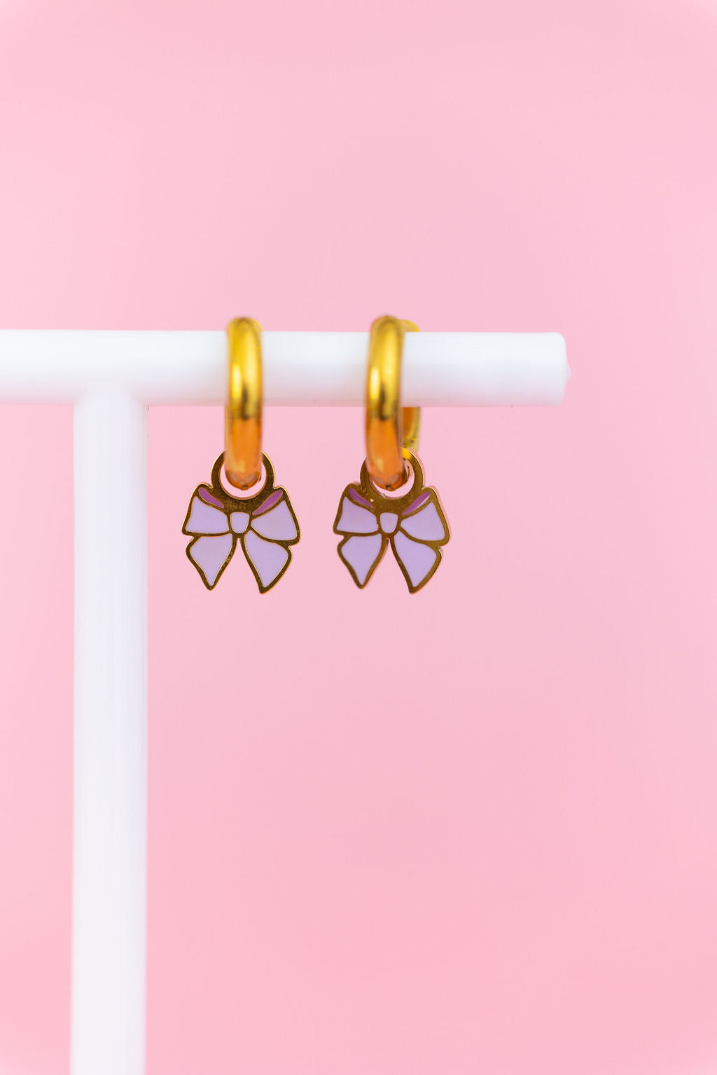 Set of lavender bow charms on gold huggie charms hanging on a white jewelry stand against a light pink background