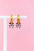 Set of lavender bow charms on gold huggie charms hanging on a white jewelry stand against a light pink background