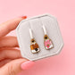 A hand with pink nails holds a small gray jewelry display card featuring two enamel dangle earrings shaped like milk cartons, one brown and one pink, against a solid pink background.