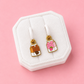 A pair of small enamel drop earrings displayed on a white jewelry stand against a pink background. One earring features a brown milk carton with a chocolate heart, and the other features a pink milk carton with a strawberry.