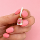 A hand with pink manicured nails holds a small enamel charm shaped like a pink milk carton with a strawberry and a white loop at the top, set against a solid pink background.