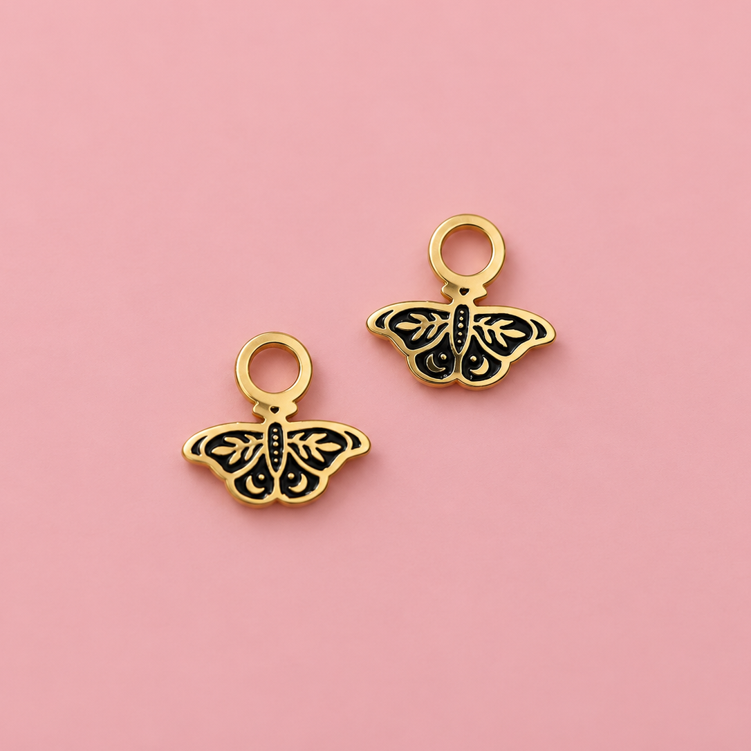 Two small butterfly-shaped jewelry charms with gold-tone metal borders and black enamel detailing, displayed on a solid pink background.
