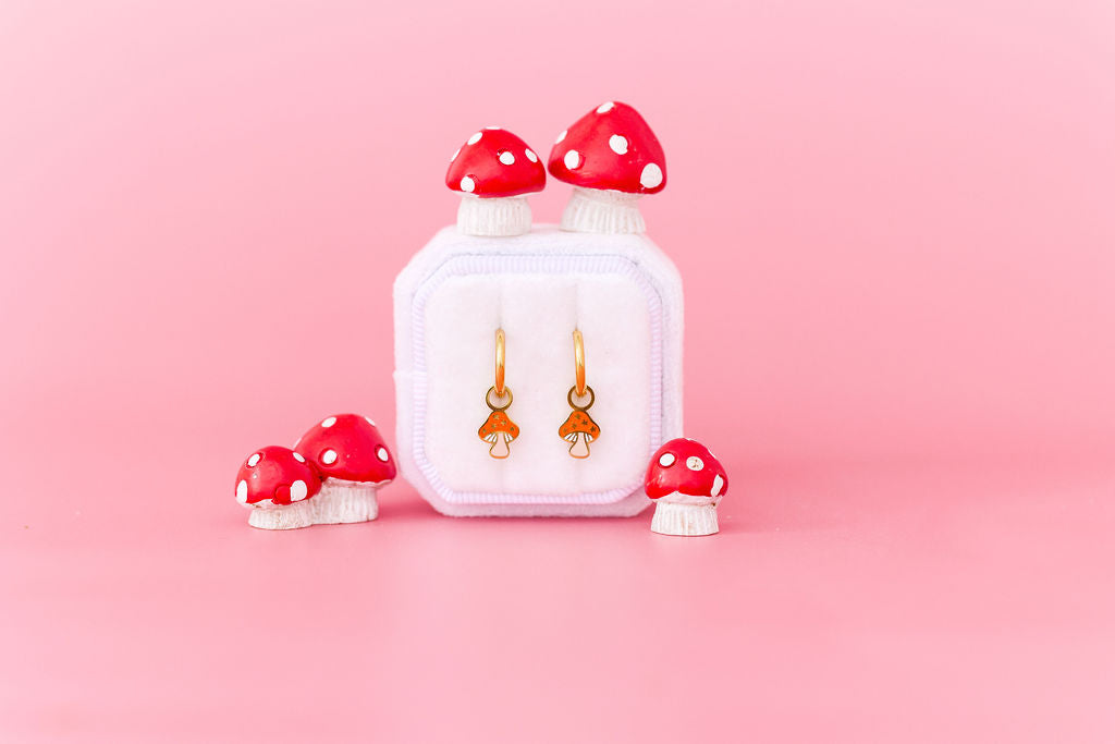 Pair of mushroom-shaped charms on 10mm gold huggie hoops in a white jewelry box with red and white polka dot mushrooms decorations on a pink background