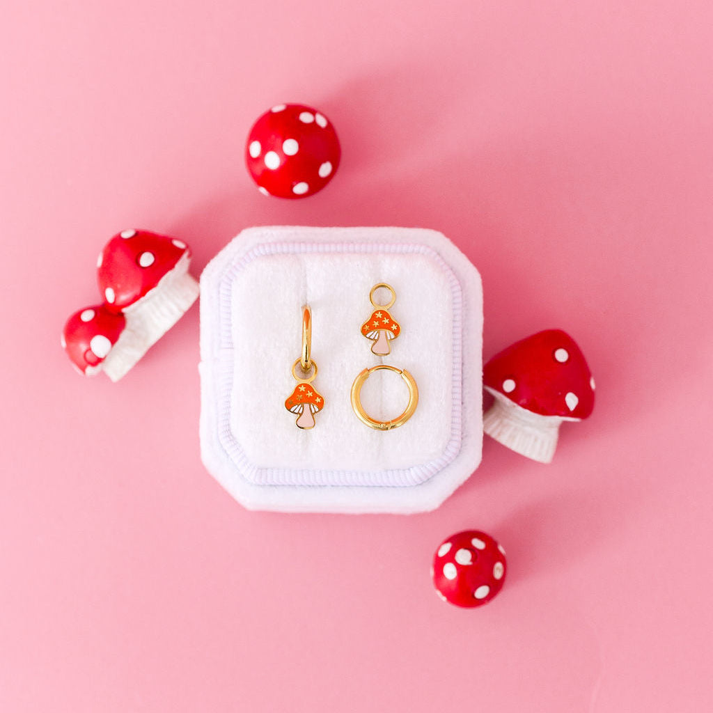 Pair of mushroom-shaped charms with 10mm gold huggie hoops in a white jewelry box with red and white polka dot mushrooms decorations on a pink background
