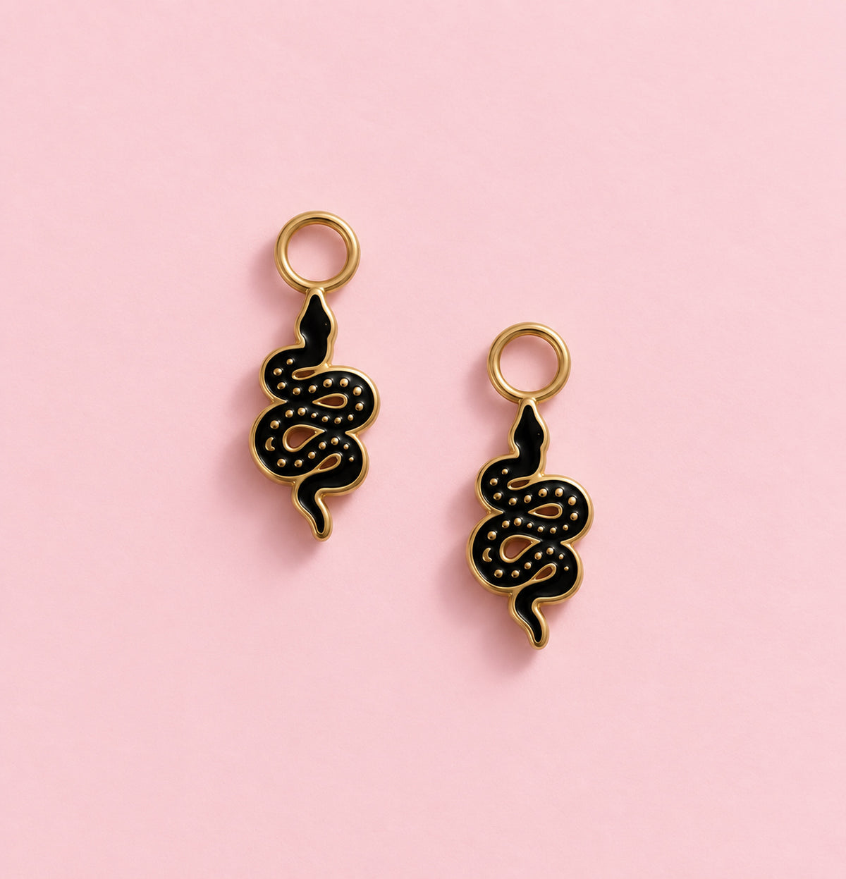 Two black and gold snake-shaped charms with gold loops at the top, displayed on a solid pink background.