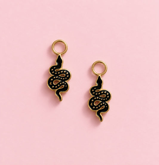 Two black and gold snake-shaped charms with gold loops at the top, displayed on a solid pink background.