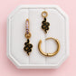 Three gold-toned earrings displayed on a white octagonal jewelry box against a soft pink background. The set includes two snake-shaped drop earrings with black enamel detailing, one with small purple stones, and one large gold hoop earring.